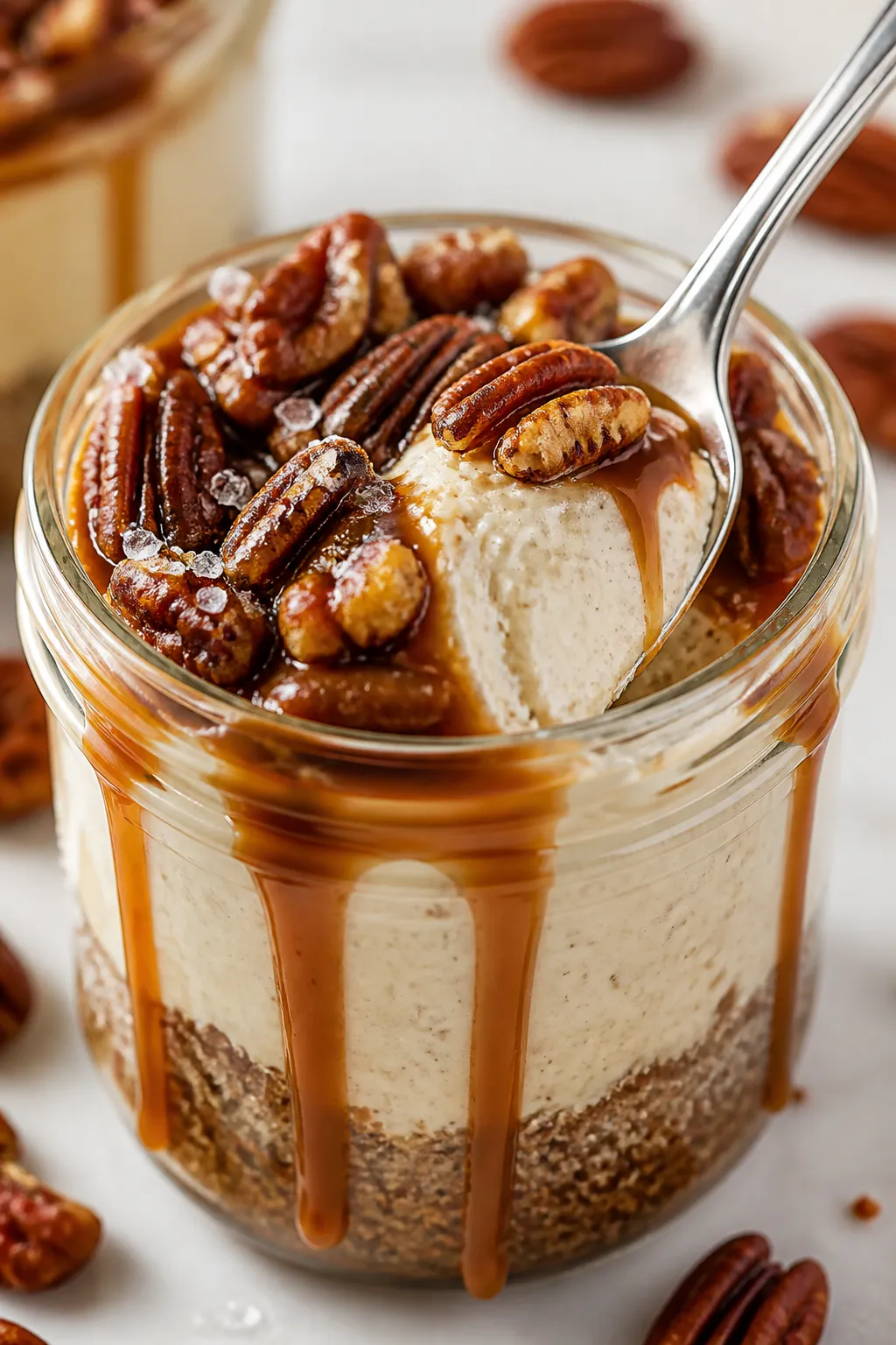 Close-up detail: A spoon sinking into a chilled caramel pecan cheesecake jar, showing thick, silky v