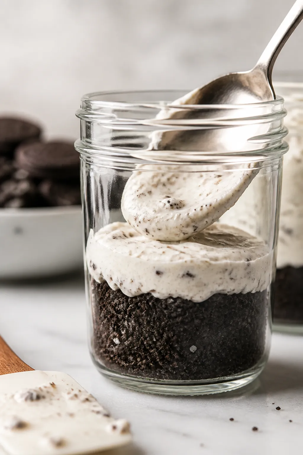 Close-up detail shot of a prepared Oreo cheesecake jar mid-assembly: a spoon gently pressing the cho